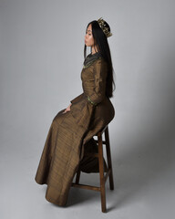 Full length  portrait of beautiful young asian woman with long hair wearing medieval fantasy gown  and gold crown costume. Graceful sitting pose,  isolated on studio background.
