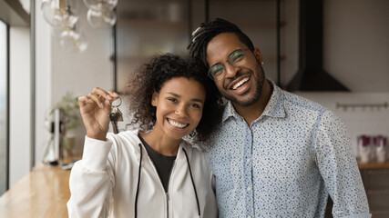 Happy homeowners smiling African married young couple standing in modern kitchen holding keys posing on camera. Moving day, own apartment, housing improvement, bank loan, affordable dwelling concept