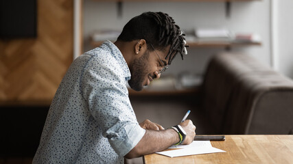 African ethnicity millennial guy sit at table fill in information survey questionnaire, making...