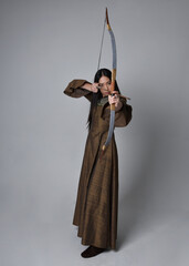 Close up  portrait of beautiful young asian woman with long hair wearing medieval fantasy gown. Graceful pose holding a long bow and arrow,  isolated on studio background.