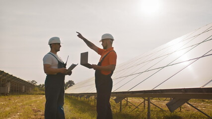 Team of industrial colleagues using tablet monitoring rows of photovoltaic solar panels at sunset. Business cooperation. Solar park. Alternative energy concept. Construction solar power plant.