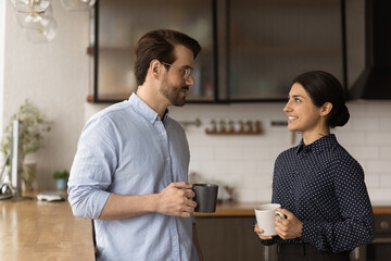 Multiracial colleagues talk during lunch break in office kitchen. Indian woman communicates with workmate hold coffee cups enjoy conversation, discuss work or personal. Good relations at work concept
