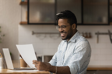 Happy African hipster man sit at table in kitchen read great news in document, got financial...