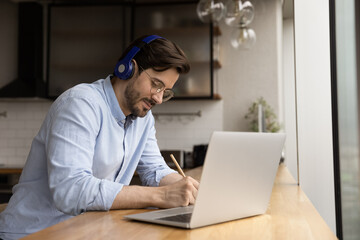 Young focused man student sit at table at home wear modern wireless headphones, listen audio course on-line, writing notes. Knowledge improvement, self-education use internet, video call event concept