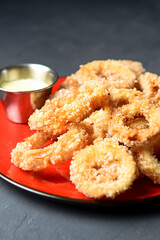 Golden onion rings and sauce in a saucepan served on a red plate. Over dark black background.