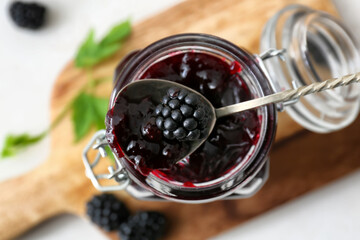 Jar and spoon with tasty blackberry jam on light background, closeup