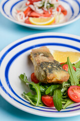 Delicious battered fish on a plate with cherry tomatoes, arugula and a piece of lemon.