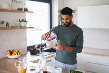 Young man preparing healthy breakfast indoors at home, pouring milk shake into glass.