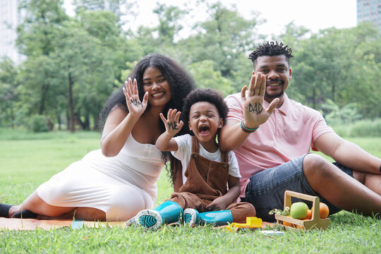 Happy African family, father mother and little son picnic and sitting on mat at park. Parents and child boy are smiling and showing messages on their palms they paint: Father, Mother, Me
