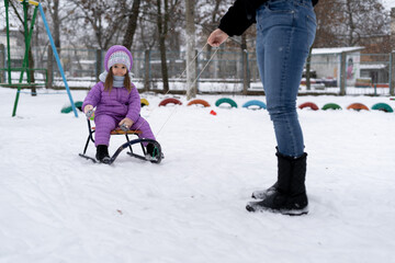 A woman is carrying a child on a sled. Mom walks with her daughter along a snowy path. Merry winter holidays. Winter fun.