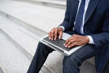 Close-up image of businessman sitting on steps and working on laptop, answering e-mails or drafting contracts