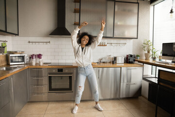 Cheerful happy pretty African woman listen music dancing alone in modern kitchen, celebrating...