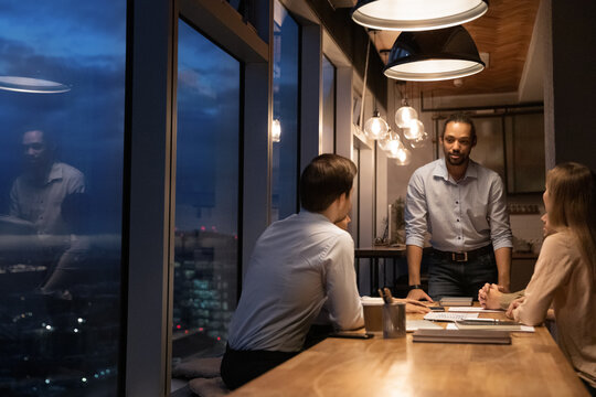 Group of young diverse colleagues entrepreneurs working until late in modern office boardroom, night city view through panoramic window. Urgency, search business solution, workshop, over hours concept