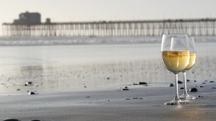Two wineglasses on sandy ocean beach. Couple of glasses and pebbles, white wine for romantic date near sea water. Cozy beachfront sunset near tide waves. Seamless looped cinemagraph. California, USA. - obrazy, fototapety, plakaty