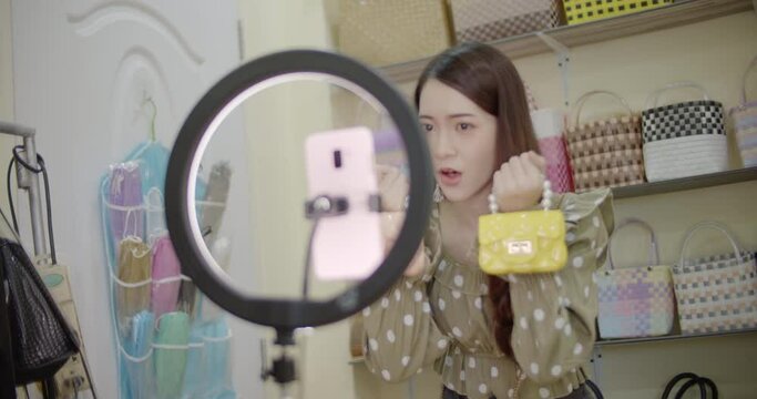 Online seller Asian woman with long brown hair is broadcasting live on mobile phone with ring light, selling women's bags in her room against the backdrop of women's bag racks.