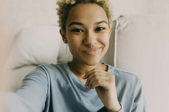 Close-up of cute African ethnicity girl recording stories for her blog using selfie camera of her smartphone, sharing information, dressed in grey blue shirt posing against cosy home background