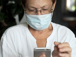 An elderly senior woman wearing a medical protective face mask handles the phone.