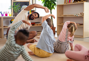 Group of small nursery school children with man teacher sitting on floor indoors in classroom,...