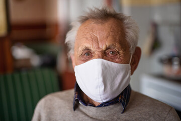 Portrait of elderly man with face mask looking at camera indoors at home, coronavirus concept.