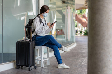 Fototapeta na wymiar Travel. woman in a white long-sleeved shirt and jeans, wearing a mask. sit on the chair and using mobile phone with suitcase placed on her right hand and backpack