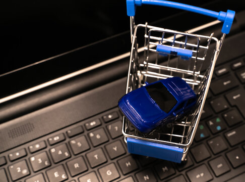 Top Down View Of A Small Toy Car In A Shopping Cart With A Laptop In The Background Illustrating Car Sales, Leasing, Rental Or Insurance. White Background With Shallow Depth Of Field.