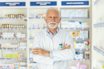 Portrait of a pharmacist in a pharmacy. Close-up shot of a male pharmacist in uniform and gray hair and beard crossed arms in the middle of the pharmacy in front of a shelf with medicines Proud worker