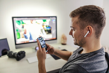 Music inspires creativity. During the break, designer or photographer listen to music and checks statuses on social media. He is sitting on a chair and has white wireless headphones in his ears