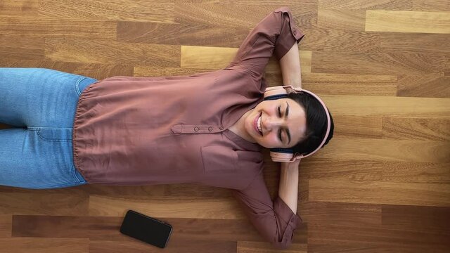 Vertical top view pretty relaxed Indian woman lying on floor listens song through wireless headphones use music digital services on cellphone. Hobby, weekend leisure, modern technology user concept