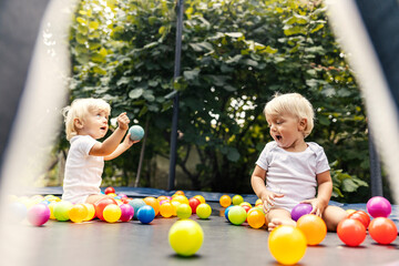 Home playroom for children. The smiling twins sit in a trampoline with a protective fence and colorful balls and play together. Sweet family moments, growing up together brother and sister - obrazy, fototapety, plakaty