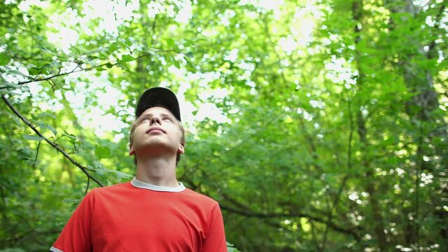 Closeup view 4k video portrait of handsome young caucasian teenage kid of 14 years old looking up in sunny sky enjoying beauty of green spring or summer nature while standing in old green wood