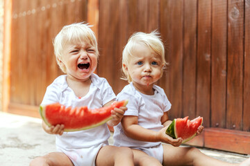 Funny photo of baby and watermelon. The twins in a white children’s ward with beautiful blue eyes and blonde hair sit on the floor of the backyard and enjoy the watermelon. Home picnic