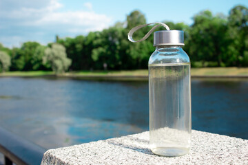 A glass bottle with water stands on a stone pedestal against the backdrop of a river and forest. It symbolizes reusable eco-friendly tableware.