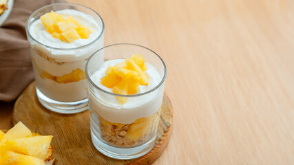 Obraz na płótnie Canvas Tasty pineapple desserts with chopped fresh juicy pineapple. Breakfast dessert with oat granola, greek yogurt and pineapple in layers in glass on wooden table. Long web banner with copy space.