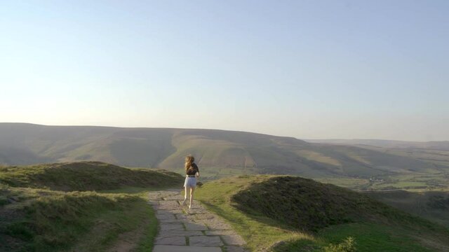Stabilised shot of young blonde woman jogging along the path on top of Mam Tor, Castleton, Peak District, England before stopping to admire the view of green rolling hills and blue skies.
