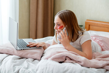 Smiling Caucasian woman in pajamas lies in bed and working using laptop. Young woman using hydrogel patches while working have conversation talk by phone