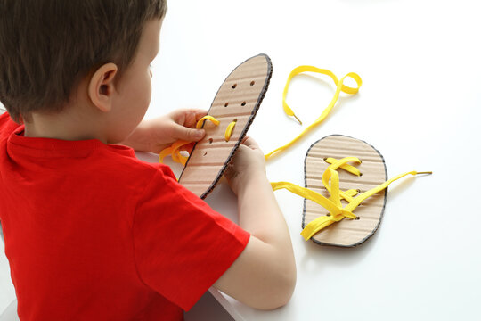 Little Boy Tying Shoe Laces Using Training Cardboard Template At White Table