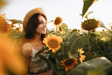 Smiling woman wearing a hat standing in a field of sunflowers in sunset