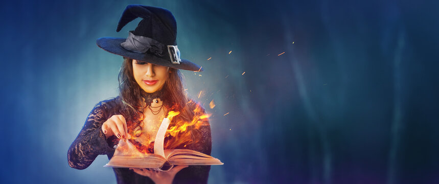 Halloween Witch girl with magic Book of spells portrait. Beautiful young woman in witches hat conjuring, making witchcraft. Over spooky dark magic forest background. Wide Halloween party art design.