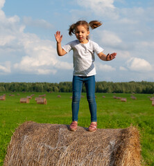 Naklejka na ściany i meble A girl in a white T-shirt with two tails on her head and a lollipop jumps on a haystack in the middle of a field against a blue sky