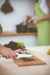 Unknown young woman slicing greens for a delicious fresh vegetarian salad while sitting and smiling at the kitchen desk, just hands, close-up. Cooking concept