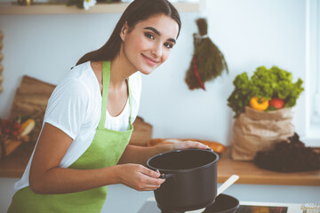 An attractive young dark-haired woman holding the dark pot of ready soup while standing and smiling near the kitchen stove. Cooking and householding concepts