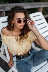 Fashion cool young curly hipster woman puts on vintage sunglasses with yellow summer blouse top and blue jeans sits and rest on the beach lounger
