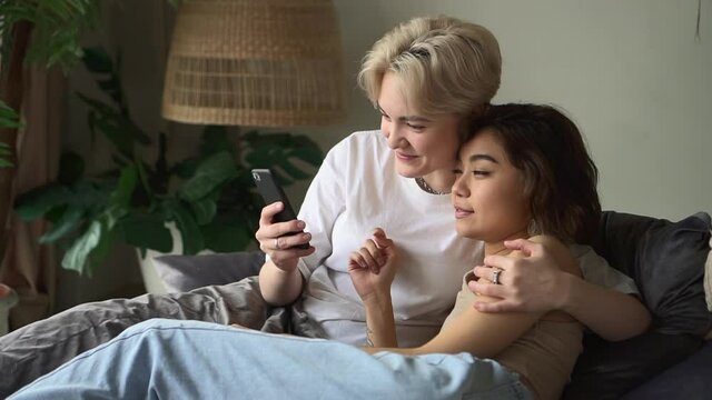 Two lesbian women using device and talking while lying on bed in home bedroom spbd.