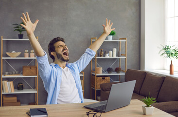 Man sitting at work table with laptop computer feeling excited, raising hands up and screaming....