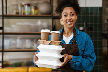 Black waitress in apron holding food boxes while working at cafe