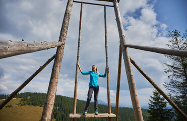 Slender blonde woman standing on seat of very large wooden frame swing, holding on to side supports and smiling happily. On the background blue sky with gray clouds, forest and mountain hills.