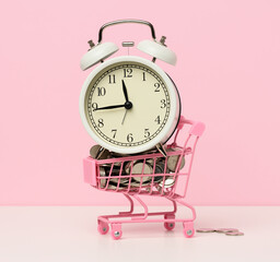 round alarm clock in a miniature shopping cart with change on a white table. Concept time is money,...