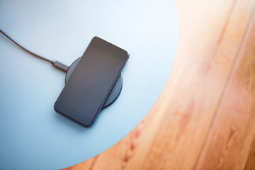 Top down view of smartphone lying on round charging pad. Light blue and wooden texture background.