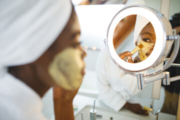 Smiling african american woman in bathroom applying beauty face mask, looking in mirror