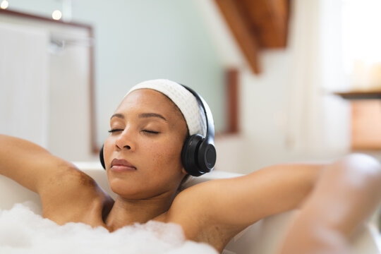 Mixed race woman in bathroom relaxing in bath wearing headphones, with eyes closed - Powered by Adobe